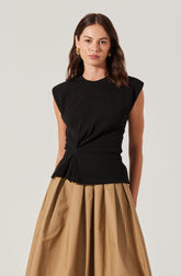 A woman is wearing a sleeveless black top with shoulder pads and a twisted detail at the waist, paired with a full-length, high-waisted skirt in a metallic beige or gold color. She is also accessorized with large gold earrings.